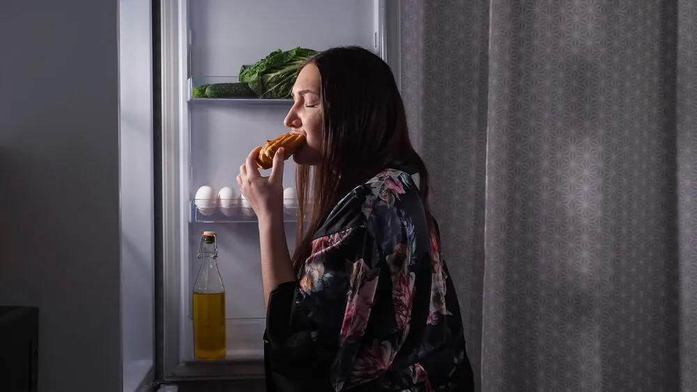 Silueta de una mujer comiendo un eclair frente a la nevera en una cocina oscura