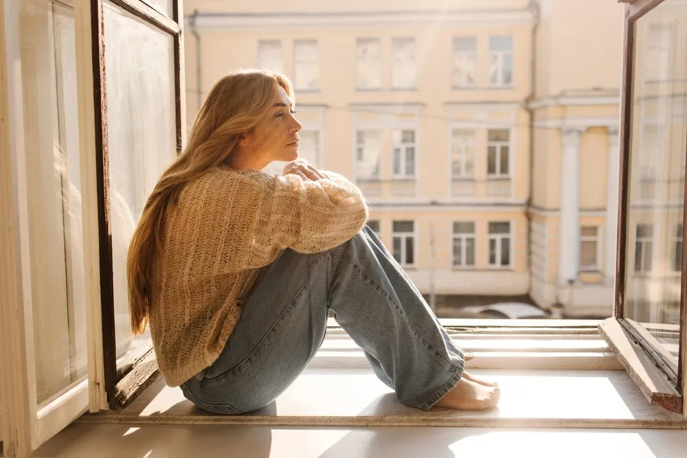 mujer adulta rubia triste de piel clara con jersey abrigado y vaqueros sentada en el alféizar de la ventana en un día soleado concepto de estado de ánimo