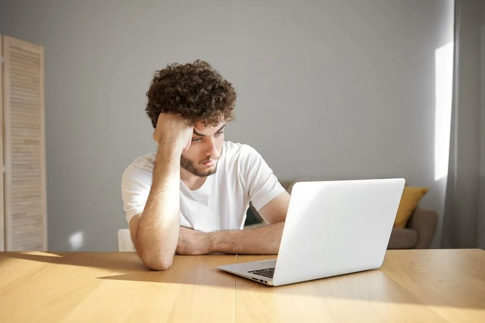 Toma horizontal de un joven frustrado y sin afeitar con camiseta blanca usando un dispositivo electrónico para teletrabajar, con aspecto cansado o aburrido mientras trabaja en un proyecto urgente temprano por la mañana.