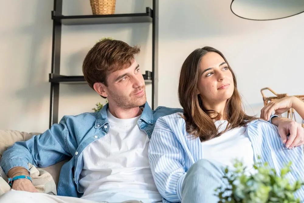Pareja joven relajándose en un sofá en su moderno salón, el hombre contempla a la mujer mientras ella mira hacia otro lado, compartiendo un momento íntimo y tranquilo de bienestar y conexión.