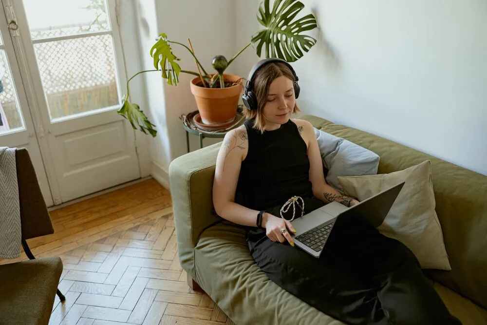 Mujer con auriculares tumbada en un sofá acogedor en casa y usando un ordenador portátil, concepto de teletrabajo.