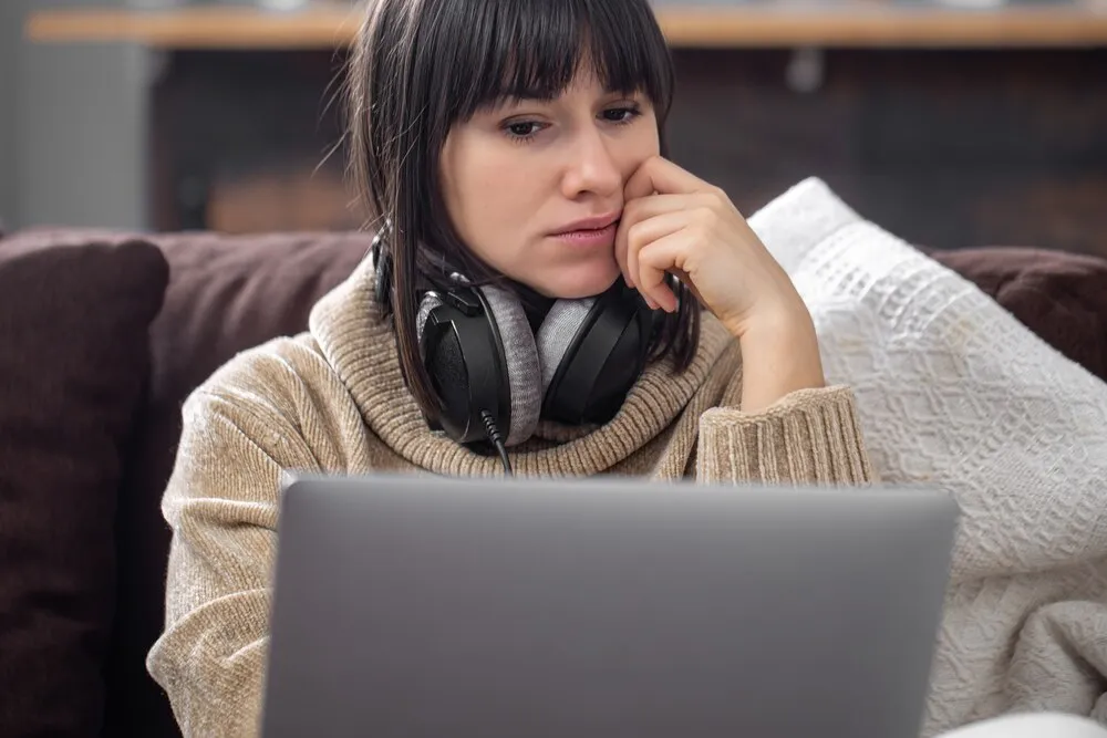 Joven morena con auriculares y un jersey acogedor mira la pantalla del portátil.
