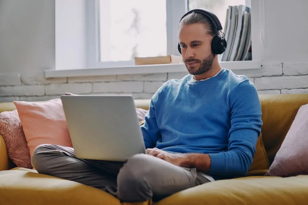 joven guapo con auriculares usando un portátil sentado en el sofá de casa
