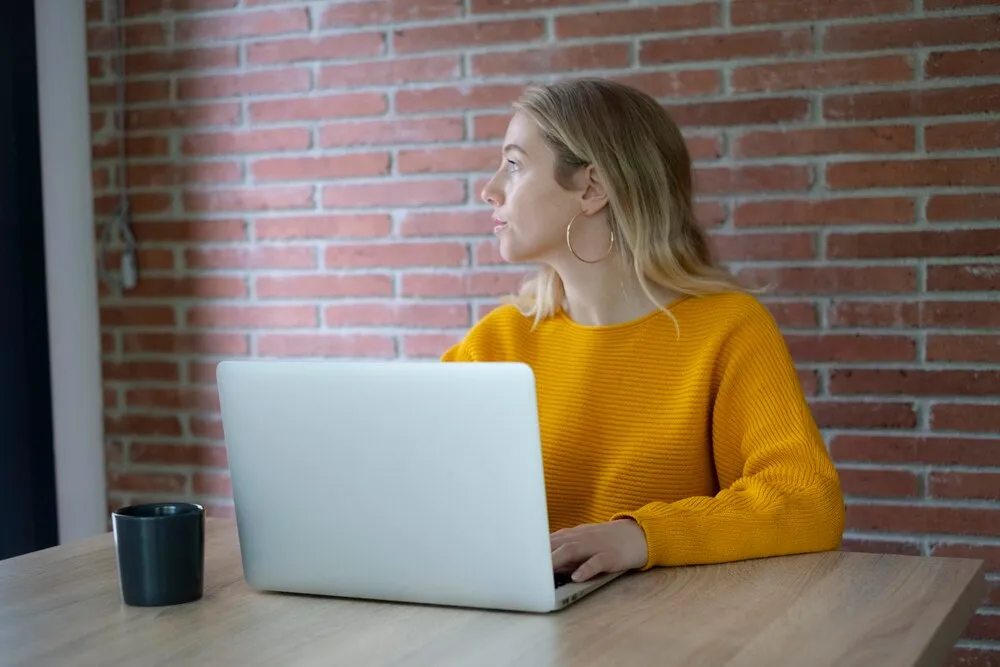 mujer con jersey amarillo trabajando en un portátil en una mesa de madera frente a una pared de ladrillo mirando por la ventana con una taza de café cerca teletrabajo pensativa y concentrada
