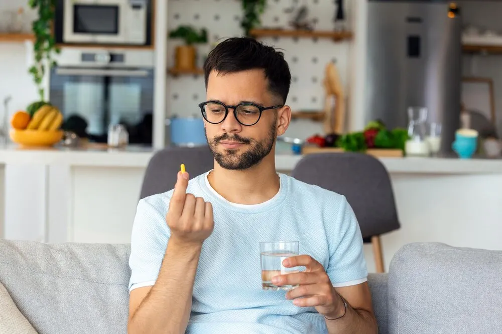 solo una pastilla puede ayudar hombre joven y guapo sosteniendo un vaso de agua y mirando una pastilla en su mano mientras está sentado en el sofá de su casa